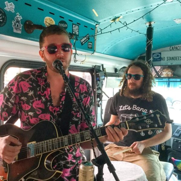 a man holding a guitar and singing into microphone with man playing drumset behind him