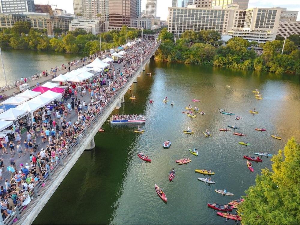 Crowded bridge festival with tents, people, and many kayaks in the river below in an urban area.