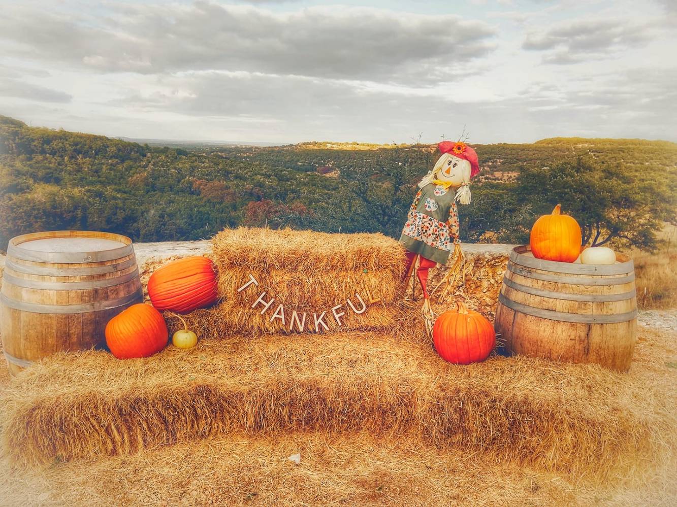 Autumn scene with hay bales, pumpkins, scarecrow, and barrels on a hillside with 'Thankful' sign.
