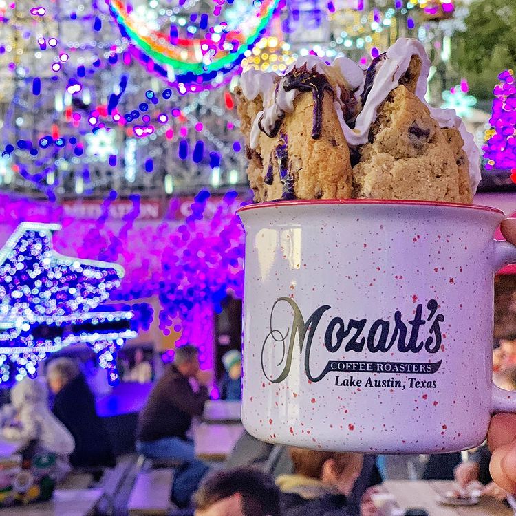 A mug with cookies, labeled 'Mozart's Coffee Roasters,' held up against a backdrop of colorful lights.