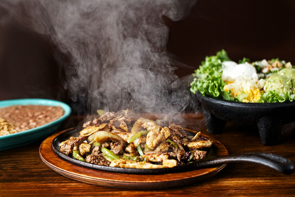Sizzling fajitas with steam, rice, beans, and salad on wooden table.