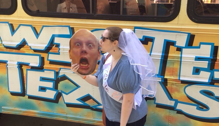 Person with bridal veil kissing a large cutout face near a graffiti-covered bus.